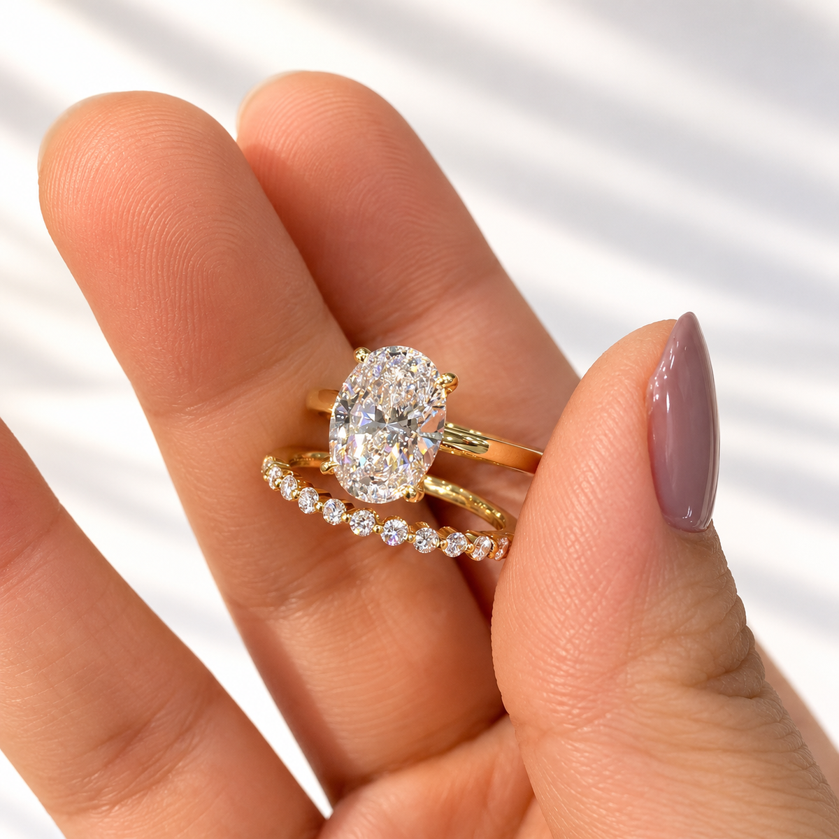 Close-up of fingers holding a yellow gold four-prong oval diamond engagement ring stacked with a delicate round diamond wedding band