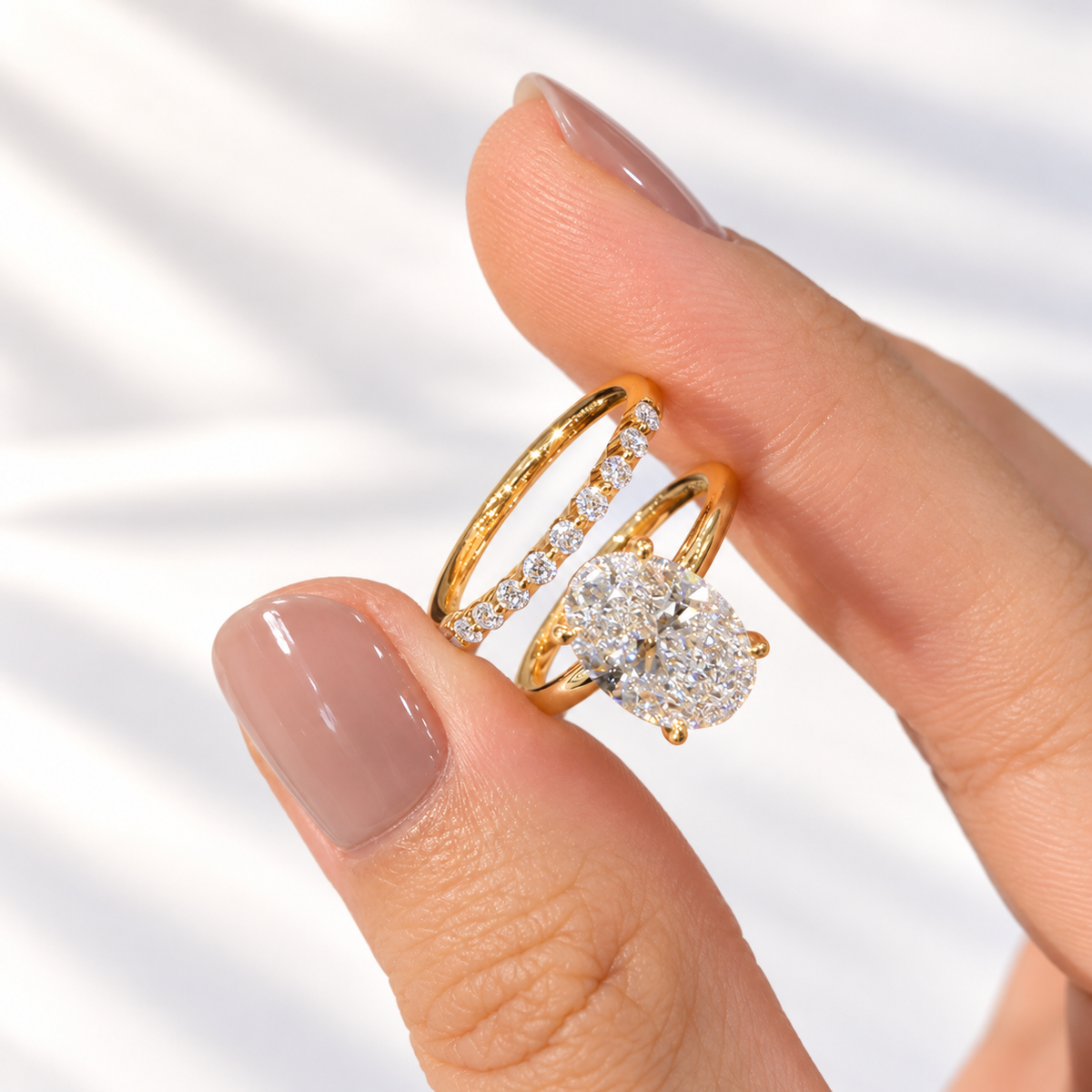 Fingers with a nude manicure holding a yellow gold oval diamond solitaire ring and a stacked wedding band of round diamonds