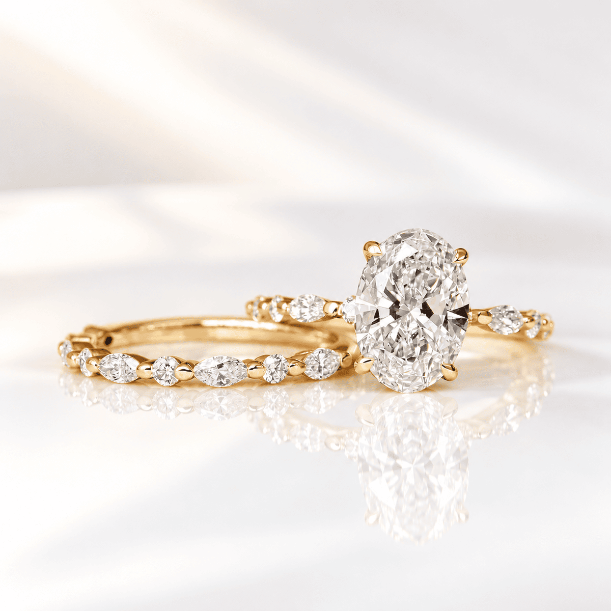 Yellow gold bridal set featuring an oval diamond engagement ring with a matching band of alternating marquise and round diamonds, shown with a soft reflection on white.