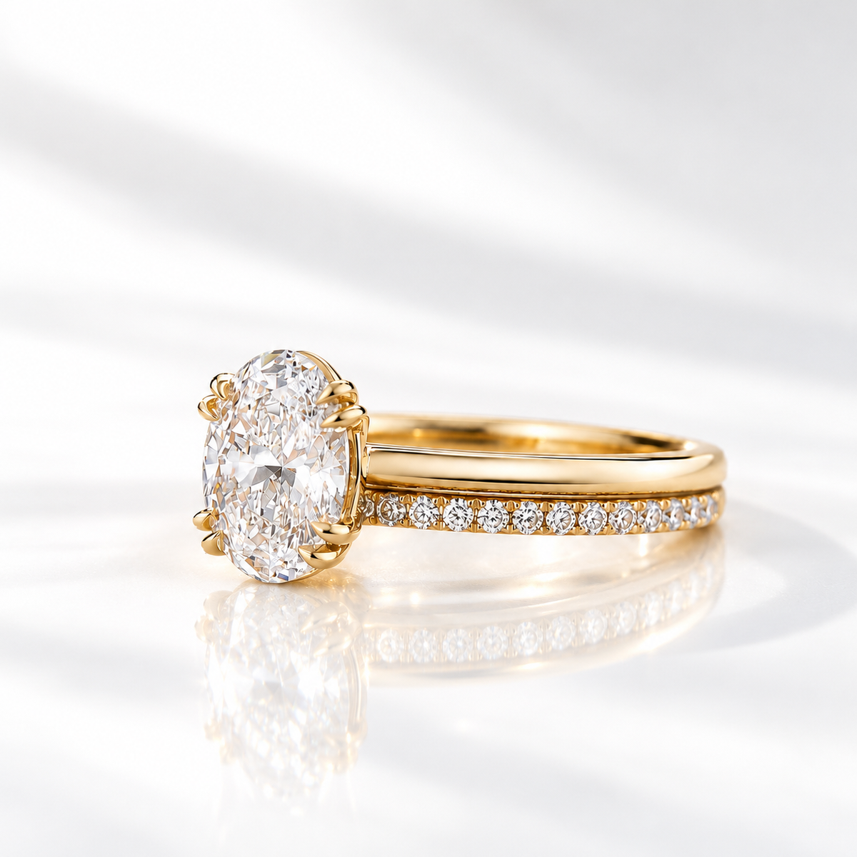 Angled side view of a yellow gold oval diamond solitaire engagement ring with double prongs, stacked with a slim pavé diamond wedding band, on a reflective white surface