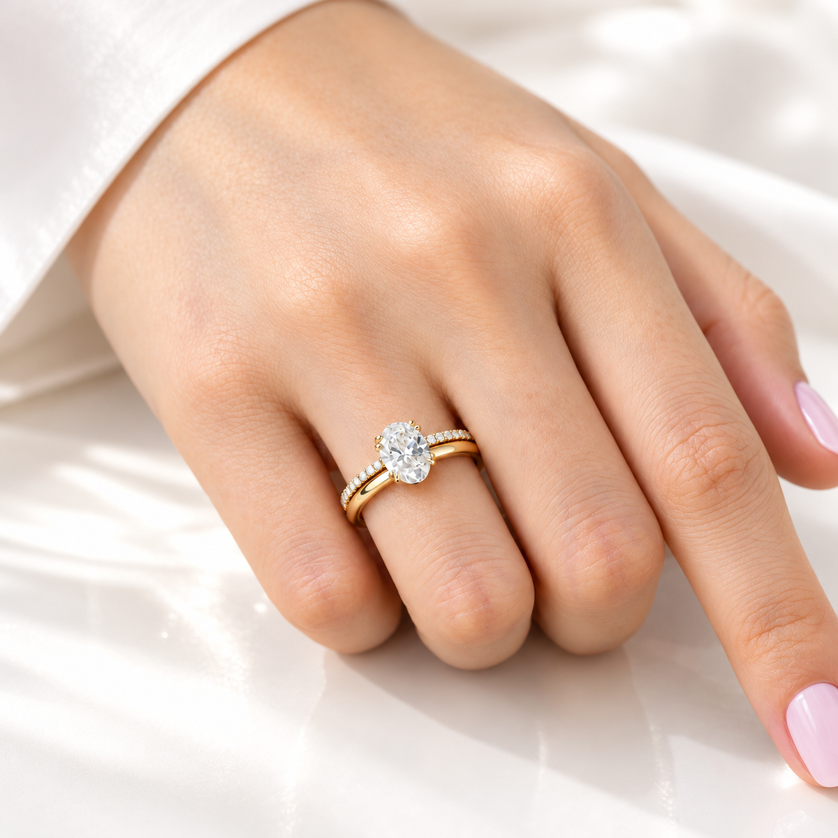 Hand with a light pink manicure wearing a yellow gold oval diamond solitaire engagement ring stacked with a slim pavé diamond wedding band, resting on white satin fabric
