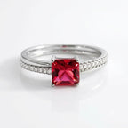 A silver ring featuring a prominent square-cut red gemstone in the center, with a pavé band set with small clear stones, shown on a reflective white surface.