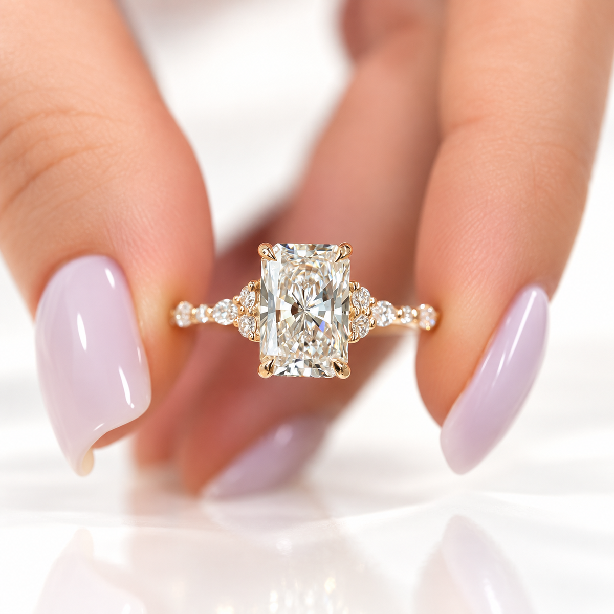 Fingers with lavender nails holding a yellow gold radiant cut diamond engagement ring with trio diamond accents and a scalloped diamond band