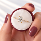 Hand with mauve nails holding a burgundy velvet ring box displaying a yellow gold radiant cut diamond engagement ring with baguette side stones along the band